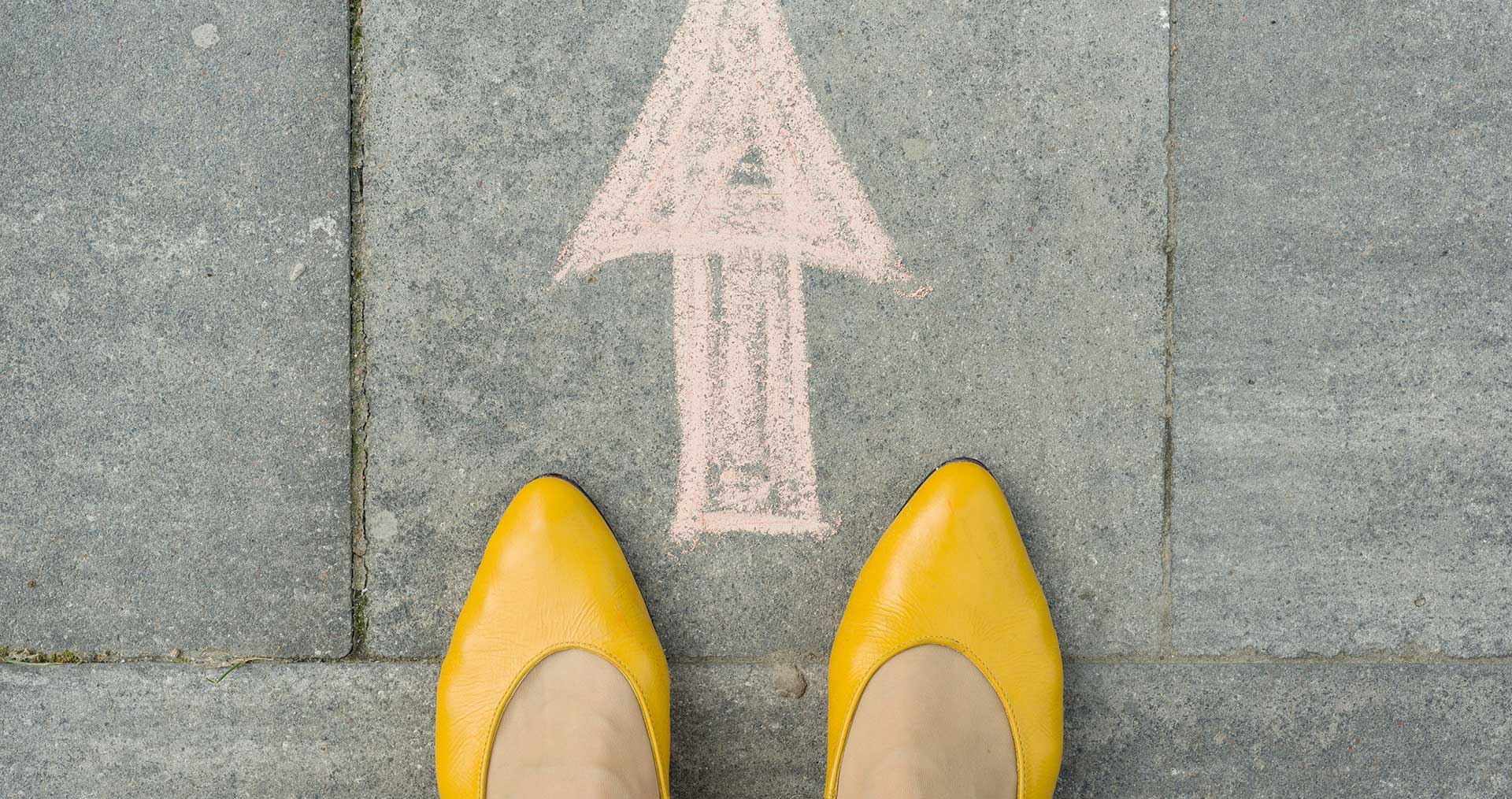 Two feet in yellow heels in front of an arrow on the sidewalk in chalk.